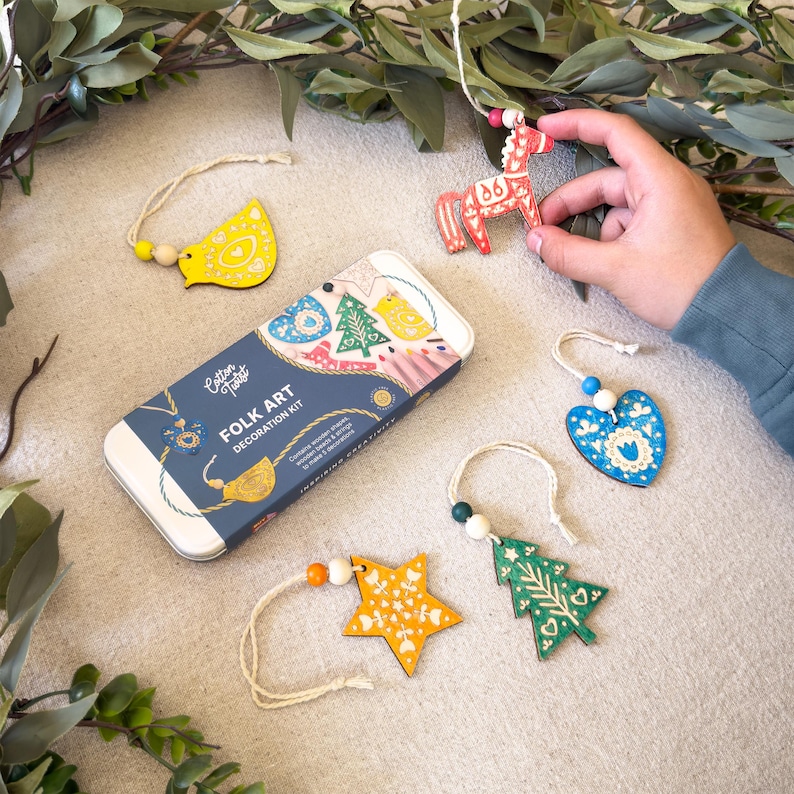 May include: A rectangular metal tin labeled Cotton Twist Folk Art Decoration Kit with five wooden ornaments including a red horse, a yellow bird, a blue heart, a yellow star, and a green tree, each featuring white painted details and hanging strings.