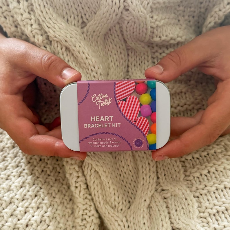 May include: A rectangular metal tin with a pink and white label that reads "Cotton Twist HEART BRACELET KIT". The tin contains colorful beads and heart-shaped beads. The kit includes wooden beads and elastic to make one bracelet.