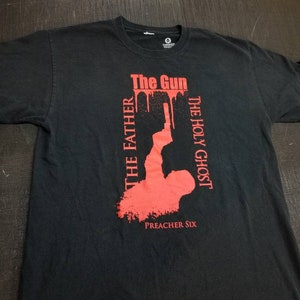 May include: Black t-shirt with a red graphic design. The graphic features a silhouette of a person holding a gun with the words "The Gun", "The Holy Ghost", and "The Father" written vertically. The text "Preacher Six" is printed below the graphic.
