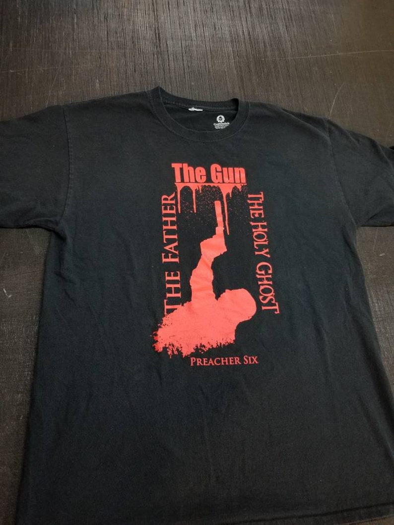 May include: Black t-shirt with a red graphic design. The graphic features a silhouette of a person holding a gun with the words "The Gun", "The Holy Ghost", and "The Father" written vertically. The text "Preacher Six" is printed below the graphic.