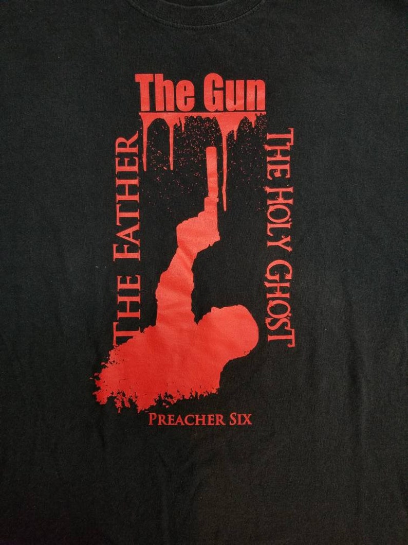 May include: Black t-shirt with red text and graphic. The text reads "The Gun", "The Holy Ghost", "The Father", and "Preacher Six". The graphic is a silhouette of a person holding a gun.