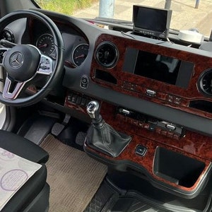 以下が含まれることがあります： 木目調のダッシュボードとメルセデス・ベンツのロゴが付いたステアリングホイールを備えた白いバン車の内装。バンは黒い革のシートと模様のある布製のシートカバーを備えています。