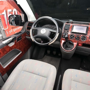 Puede incluir: Vista interior de una furgoneta Volkswagen. El salpicadero y los paneles de las puertas presentan un acabado de veta de madera. El volante es negro con un emblema plateado. Los asientos son de color gris claro con un diseño estampado.