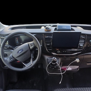 May include: Interior view of a Ford Transit van dashboard. The black dashboard features a steering wheel with the Ford logo, a large infotainment screen, and various controls. A charging cable is plugged into a USB port.