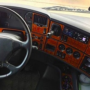 May include: Interior of a Scania truck cab with a black steering wheel and wood-effect dashboard. The dashboard includes various controls, gauges, and a radio. A small rearview mirror is visible above the windscreen.