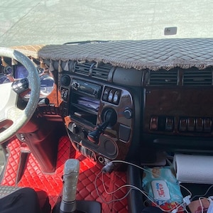 May include: The interior of a truck cab with a brown wood-grain dashboard, a white steering wheel with a black logo, and red quilted floor mats.