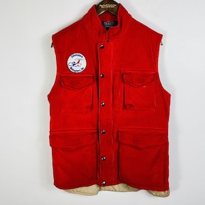 May include: A vibrant red corduroy vest with several pockets and a button-up front. The vest has a patch with the text "Commodore Helicopters, Inc." and the brand name "GANT" on the collar.