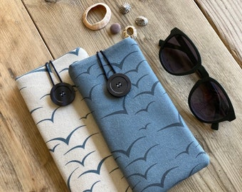 May include: Two fabric eyeglass cases with a seagull pattern. The case on the left is beige with dark blue seagulls. The case on the right is blue with white seagulls. Both cases have a black button closure.