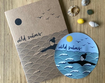 May include: A brown notebook with the text "wild swims" and a black and white illustration of a person swimming in the ocean. A blue and white sticker with the text "wild swims" and a black and white illustration of a person swimming in the ocean is attached to the notebook.