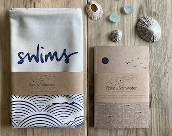 May include: A white tea towel with a blue wave pattern and the word "swims" printed in blue. The tea towel is packaged with a brown paper label that says "Rock & Saltwater" and "Screen printed tea towel".