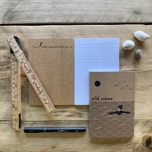 May include: A wooden ruler, black pen, and several notebooks are arranged on a wooden surface. One notebook is open, revealing lined paper, while another features the text "wild swims" and a swimmer illustration. The text "creative adventures start here" is also visible.