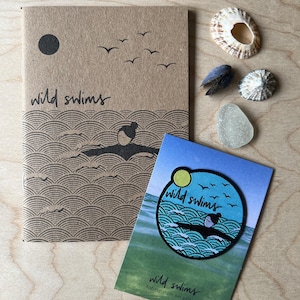 May include: A tan notebook with a black graphic of a person swimming in waves, a sun, and birds, with the text "wild swims" printed on the cover. Next to it is a blue circular patch with a matching design and a card labelled "wild swims".