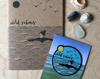 May include: A tan notebook with a black graphic of a person swimming in waves, a sun, and birds, with the text "wild swims" printed on the cover. Next to it is a blue circular patch with a matching design and a card labelled "wild swims".