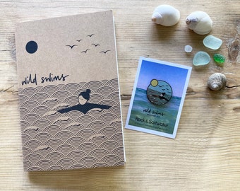 May include: A brown notebook with the words "wild swims" printed on the front. The notebook has a black circle and a graphic of a person swimming in the ocean. A small enamel pin with the words "wild swims enamel pin" and "Rock & Saltwater" is shown on a white card.