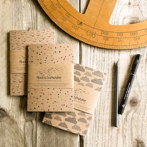 May include: Three brown paper notebooks with different designs. The first notebook has a pattern of birds, the second has a colourful confetti pattern, and the third has a cloud pattern. Each notebook has a label with the text "Rock & Saltwater" and a website address.