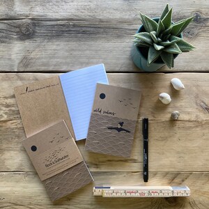 May include: A collection of notebooks with brown covers, some with the text "wild swims" and "Rock & Saltwater". A black pen and a folding ruler are also present. A small potted succulent and seashells are on a wooden surface.