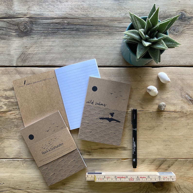 May include: A collection of notebooks with brown covers, some with the text "wild swims" and "Rock & Saltwater". A black pen and a folding ruler are also present. A small potted succulent and seashells are on a wooden surface.