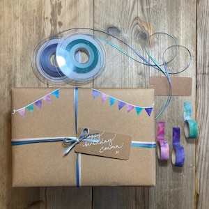 May include: A wrapped gift in brown paper, adorned with blue and teal ribbons and watercolor pennants. The gift is decorated with washi tape and a tag that reads "happy birthday Emma". Two spools of ribbon and blank tags are also visible.