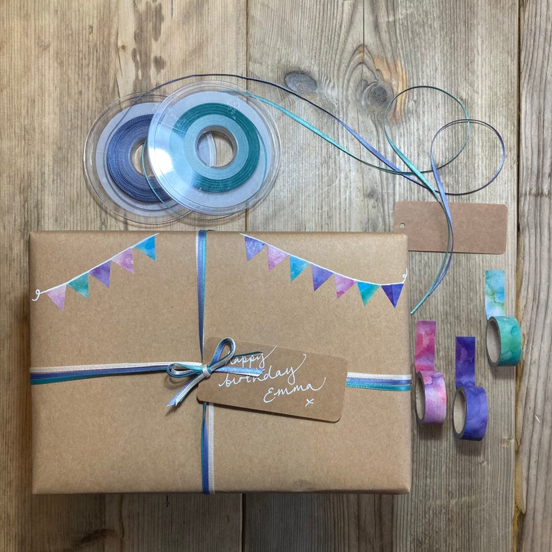 May include: A wrapped gift in brown paper, adorned with blue and teal ribbons and watercolor pennants. The gift is decorated with washi tape and a tag that reads "happy birthday Emma". Two spools of ribbon and blank tags are also visible.