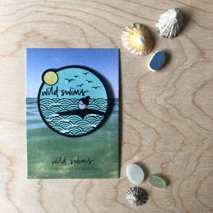 May include: A circular fabric iron-on patch featuring a graphic of a person swimming in ocean waves under a yellow sun and birds, with the text "wild swims". The patch is mounted on a rectangular card labelled "wild swims fabric iron on patch".