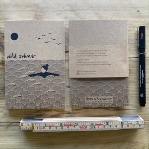 May include: Two brown notebooks with the words "wild swims" and a swimmer illustration. A black pen and a ruler are next to the notebooks. The notebooks have a wave pattern design.