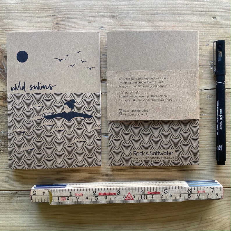 May include: Two brown notebooks with the words "wild swims" and a swimmer illustration. A black pen and a ruler are next to the notebooks. The notebooks have a wave pattern design.