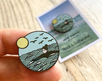May include: A blue and green enamel pin featuring a person swimming in the ocean with a yellow sun and birds flying overhead. The pin says "wild waves" and "Rock & Saltwater".