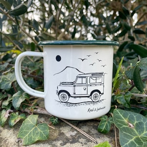 May include: White enamel mug with a green rim. The mug features a black and white illustration of a Land Rover driving through a mountainous landscape. The text "Rock & Saltwater" is printed on the bottom of the mug.