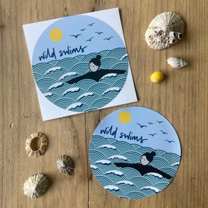 May include: Two blue and white stickers with the text "wild swims" and a silhouette of a person swimming in waves. The stickers are on a wooden surface.