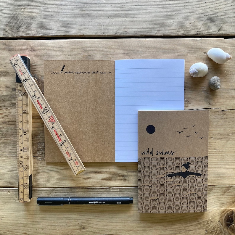 May include: A wooden ruler, black pen, and several notebooks are arranged on a wooden surface. One notebook is open, revealing lined paper, while another features the text "wild swims" and a swimmer illustration. The text "creative adventures start here" is also visible.