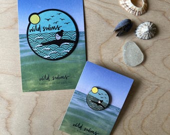 May include: A circular fabric iron-on patch and a circular enamel pin, both featuring a graphic of a person swimming in ocean waves under a yellow sun and birds, with the text "wild swims" printed on their respective display cards.