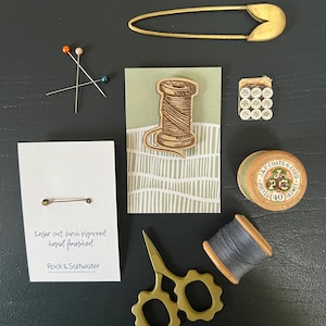 May include: A selection of haberdashery items, including a large gold safety pin, wooden thread spools, pins, buttons, and a laser-cut birch plywood pin with text. The items are arranged on a dark surface.