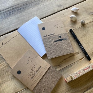 May include: A collection of kraft paper notebooks with lined pages, featuring illustrations and text. One notebook reads "wild swims" with a swimmer graphic. Another reads "Rock & Saltwater". A black pen and a ruler are also present.