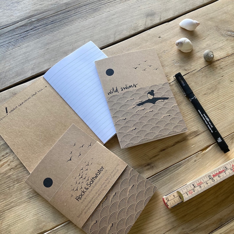 May include: A collection of kraft paper notebooks with lined pages, featuring illustrations and text. One notebook reads "wild swims" with a swimmer graphic. Another reads "Rock & Saltwater". A black pen and a ruler are also present.