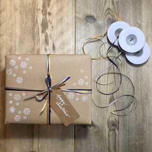 May include: A wrapped gift with a kraft paper exterior, adorned with white snowflake patterns and tied with navy, silver, and gold ribbons. A small tag reads "Happy Christmas". Rolls of matching ribbon are nearby.
