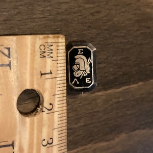 May include: A black and gold enamel pin with a Greek helmet and the letters "ΣΕ" on a black background.