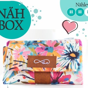 Könnte beinhalten: Eine weiße Stofftasche mit einem Blumendruck in Rosa-, Blau- und Gelbtönen. Die Tasche hat eine braune Lederklappe mit einem Unendlichkeitszeichen und dem Wort "love" darauf. Die Tasche ist mit "NÄH BOX" und "Nählevel" sowie drei Knöpfen beschriftet.