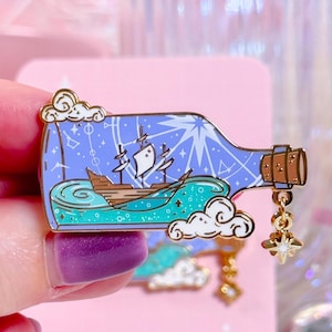 Puede incluir: Pin de esmalte dorado con un barco en una botella con un cielo estrellado azul y morado, olas verdes y nubes blancas. La botella tiene un tapón de corcho y una cadena dorada con un dije de estrella.