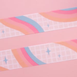 May include: Two rolls of washi tape with a rainbow and grid pattern on a pink background. The tape has a light blue, pink, and orange colour scheme with white stars.