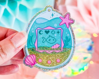 May include: A blue and pink glitter sticker of a Tamagotchi-like pet with a fish inside. The sticker has a seashell and a starfish on it. The screen of the pet reads "C O O T".