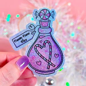 May include: A holographic sticker featuring a purple potion bottle design with a candy cane heart and a lollipop on top. The sticker has the text "Candy Cane Fizz" on a tag. The background is a soft pink with bokeh lights.