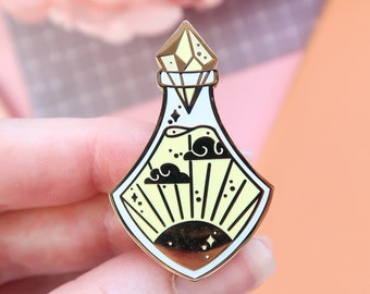 May include: Gold-tone enamel pin featuring a potion bottle with a crystal stopper. The bottle contains a yellow liquid with a sun and clouds design.