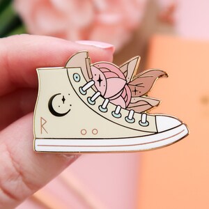 May include: A gold-tone enamel pin of a cream-colored high-top sneaker with a pink flower and leaves inside. The sneaker has a crescent moon and stars on the side and the word "Roo" on the sole.