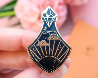 May include: Gold and black enamel pin of a potion bottle with a diamond stopper. The bottle contains a sun, clouds, and stars.