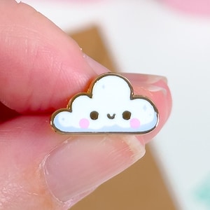 May include: A small, white cloud-shaped enamel pin with a gold border. The cloud has a smiling face with pink cheeks and black eyes. The pin is held between fingers against a blurred background.