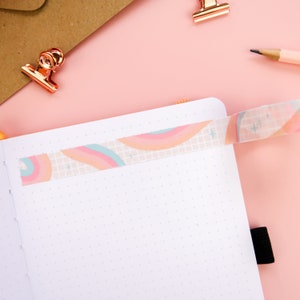 May include: A white notebook with a dotted grid pattern and a rainbow patterned washi tape across the top. The notebook is open and lying flat on a pink surface.