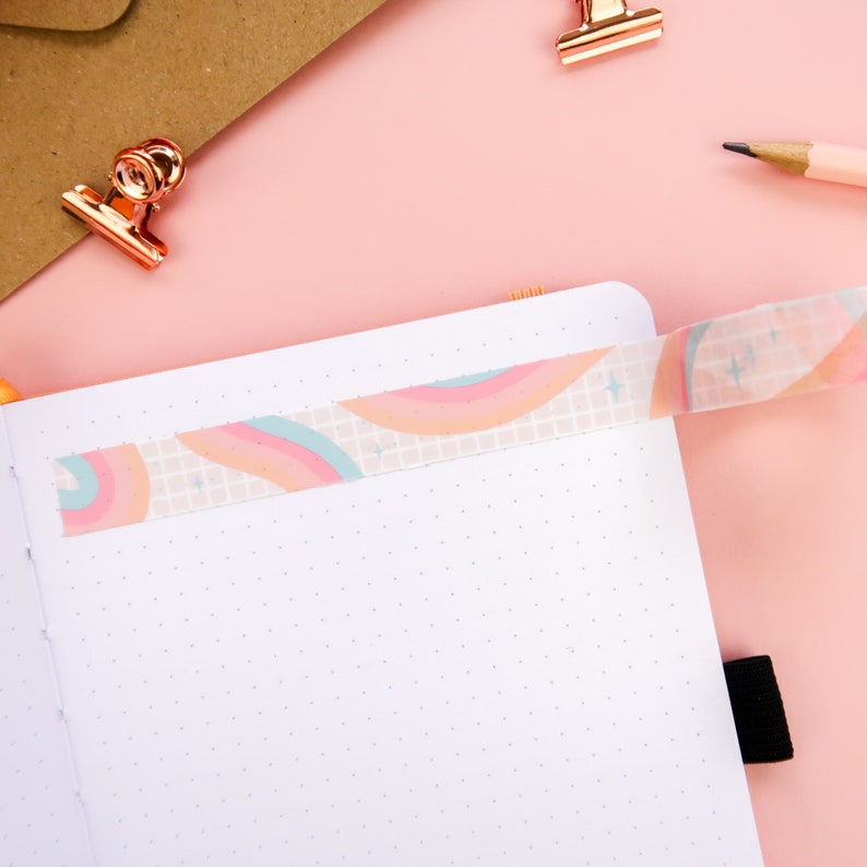May include: A white notebook with a dotted grid pattern and a rainbow patterned washi tape across the top. The notebook is open and lying flat on a pink surface.