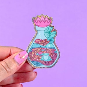 May include: A clear glass bottle with a pink flower on top, filled with glitter, water, and three green frogs. The bottle is a sticker with a shiny, glittery finish.