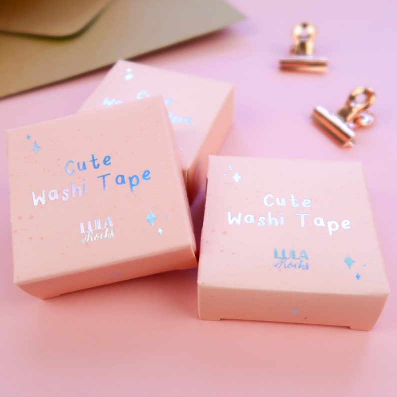 May include: Three pink boxes with the text "Cute Washi Tape" and the logo "LULA Rocks" in a light blue color. The boxes are on a pink surface.