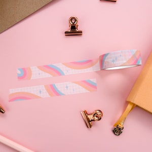 May include: Two rolls of washi tape with a rainbow and star pattern on a pink background. The tape is white with a grid pattern and features pink, blue, and orange rainbows with white stars.